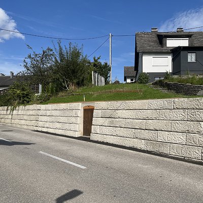 Stützmauer aus Betonblöcken sichert Hang am Straßenrand – mit integriertem Zugang zum Mostkeller